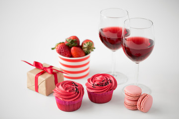 valentines day and sweets concept - close up of frosted cupcakes, macarons, strawberries, glasses of red wine and gift box on white background