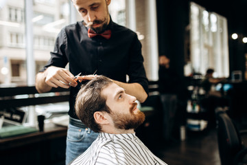 Mustachioed barber dressed in a black shirt with a red bow tie makes a stylish hairstyle to young man in a barbershop