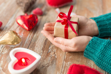 christmas, valentines day and holidays concept - close up of female hands holding gift box with heart shaped decorations and candle burning on wooden background