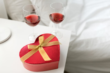 valentines day and romantic concept - heart shaped gift box and two glasses of red wine on bedroom nightstand at home