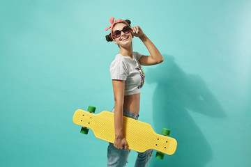 Funny young girl in sunglasses and pink bow on her head dressed in jeans and top stands with yellow skateboard on the back on the blue background in the studio