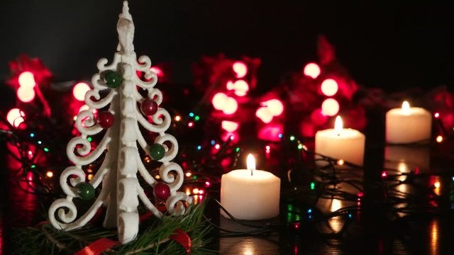 Small decorative white Christmas tree surrounded by three burning white candles on the background of flickering Christmas lights, black background