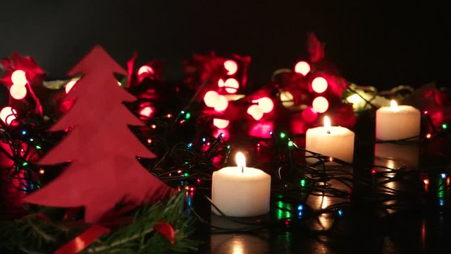 Small decorative red Christmas tree surrounded by three burning white candles on the background of flickering Christmas lights, black background