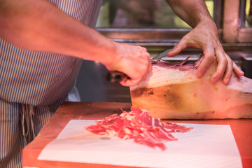 A butcher is seen cutting jamon meat with a butcher knife. The jamon looks fresh and is ready to be sold after being cut. He is seen wearing a stripe Apron.