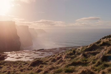 Cliff of Moher  on sunny day, sun flare, haze, clouds,  warm orange tones, county Clare, Ireland.