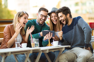 Group of four friends having a coffee together. Two women and two men at cafe talking laughing and enjoying their time using digital tablet.