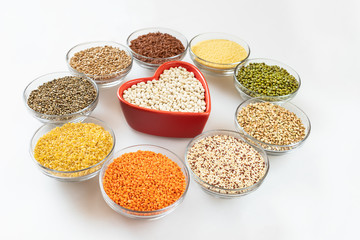 Round glass bowls with superfood are standing around red heart-shaped bowl on white background.