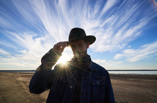 handsome bearded male traveler looking to camera and touching his felt hat with a beautiful sunset sky in background - Powered by Adobe