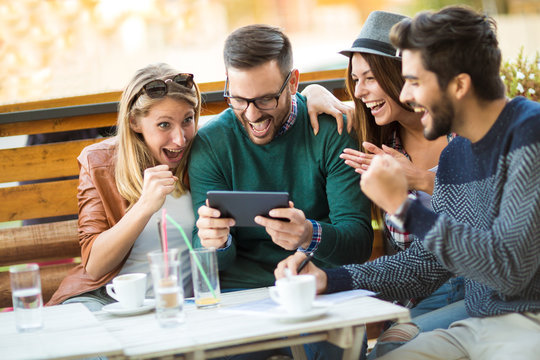Group of four friends having a coffee together. Two women and two men at cafe talking laughing and enjoying their time using digital tablet.