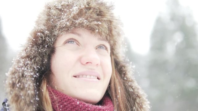 A young woman in a warm winter hat with earflaps looks at the falling snow. Close-up, slow motion. Siberian frosts and warm clothes in winter.