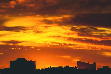 Cityscape with vivid fiery dawn. Amazing warm dramatic cloudy sky above dark silhouettes of city buildings. Orange sunlight. Atmospheric background of sunrise in overcast weather. Copy space.