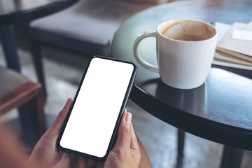 Mockup image of woman's hands holding and using a black mobile phone with blank screen with coffee cup on table