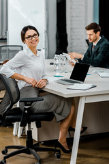 selective focus of smiling businesswoman in eyeglasses looking at camera in office with multiracial colleagues