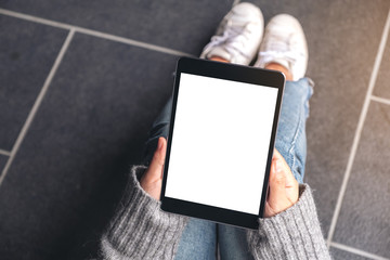 Top view mockup image of woman's hands holding and using black tablet pc with blank white desktop screen while sitting on the floor