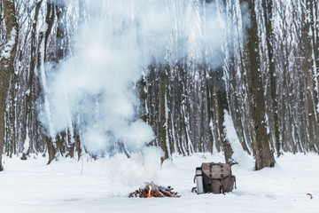 Campfire burns in the snow in the woods, on a background of snow covered trees. fire burning in cold winter. Snow, forest. Tourism. Flames, backpack and a thermos with coffee, tea