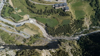Aerial photo of the Pyrenean mountains in the summer. There are green mountains with trees and rocks, ring roads with cars down below at Andorra La Vella, Andorra. There are city with brown buildings