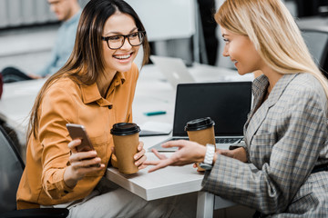 cheerful businesswomen using smartphone together at workplace in office