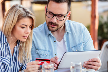 Beautiful young couple shopping online while sitting in a cafe. Man is holding a tablet and entering credit card details