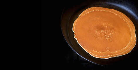 Fried pancake in a frying pan. Black background.  Hot ruddy pancake in the pan. Place for text