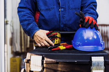 Electrian engineer holding multimeter and tools in hand, standing behind the heavy duty tool box, image including power cord, Blue hard hat (helmet) and gloves.