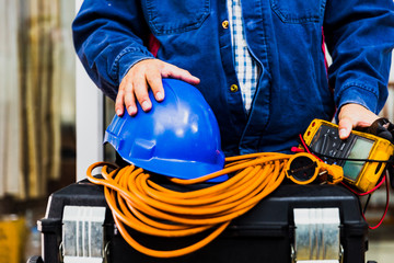 Electrian engineer holding multimeter and tools in hand, standing behind the heavy duty tool box, image including power cord, Blue hard hat (helmet) and gloves. - obrazy, fototapety, plakaty