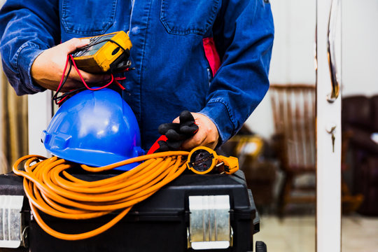 Electrian engineer holding multimeter and tools in hand, standing behind the heavy duty tool box, image including power cord, Blue hard hat (helmet) and gloves.