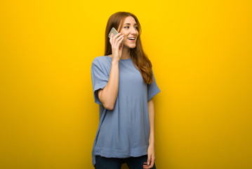 Young redhead girl over yellow wall background keeping a conversation with the mobile phone