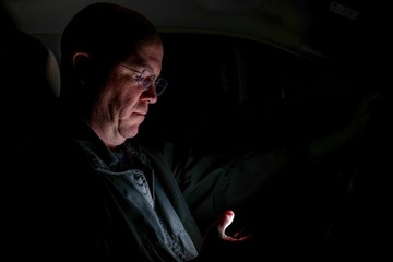A caucasion man pretends to engage in the reckless act of texting while driving a vehicle, with the glow of the screen on his face in the dark.