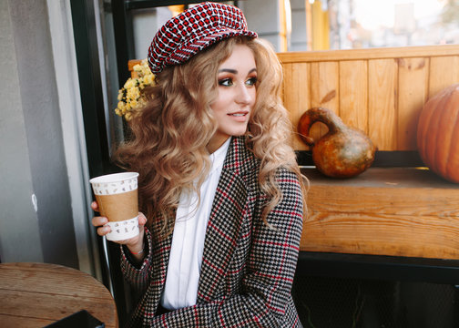 Autumn luxury look, pretty cool smiling young woman with coffee cup using smartphone walking in city, happy female model wearing elegant hat, brown jacket outdoors over outdoor cafe  background