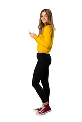 A full-length shot of a young girl with yellow sweater sending a message with the mobile on isolated white background