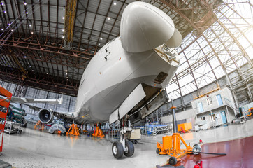 Passenger airplane on maintenance of engine and fuselage check repair in airport hangar. With an open hood on the nose under the cockpit of pilots.