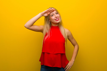 Young girl with red dress over yellow wall saluting with hand