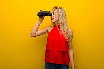 Young girl with red dress over yellow wall and looking in the distance with binoculars