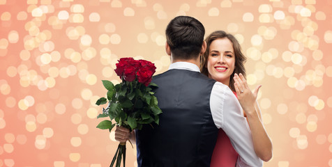 love, couple, proposal and people concept - happy woman with engagement ring and bunch of roses hugging man over living coral background and festive lights