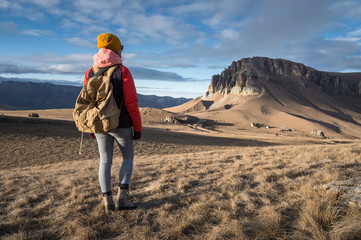 Portrait from the back of a girl traveler in a jacket with a cap and a backpack stands on the background of an epic landscape with rocks