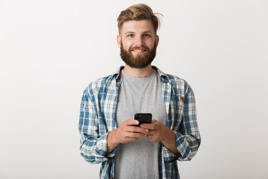 Handsome young bearded man standing isolated over white wall background using mobile phone.