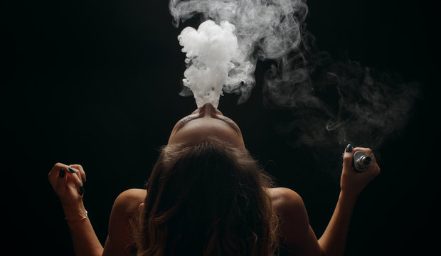 Redhead woman vaping electronic cigarette with smoke on black background closeup. Young woman smoking e-cigarette to quit tobacco. Vapor and alternative nicotine free smoking concept, copy space 