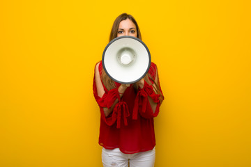 Young girl with red dress over yellow wall shouting through a megaphone to announce something