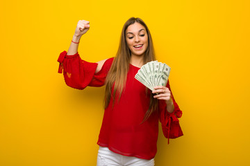 Young girl with red dress over yellow wall taking a lot of money