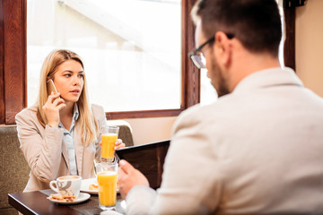 Two young male and female business partners having a meeting over breakfast in a cafe. Woman is talking on the phone and drinking orange juice