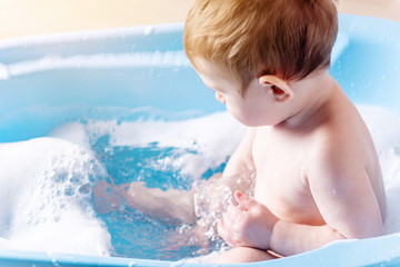 Fototapeta na wymiar Cute toddler boy washing in blue bath in bathroom. Child plays with water and soap foam.
