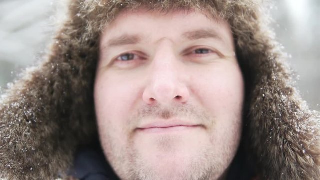 A man in a warm hat with earflaps stands in a winter park, snow is falling. Close-up of his face, he is looking at the camera and smiling. His fat face gets bigger