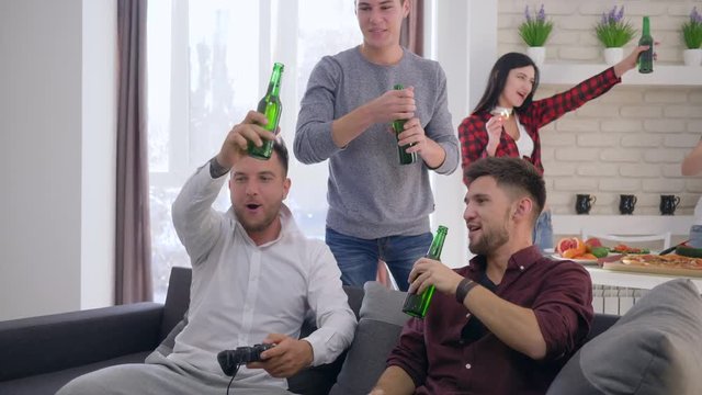joyful friends toasting beer bottles and playing video games sitting on sofa on backgrounf of company of girlfriends at home party