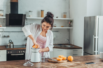 smiling girl making fresh orange juice with juicer at wooden table in kitchen