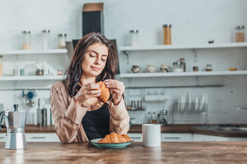 young beautiful woman having breakfast with croissants and coffee at wooden table in kitchen at home