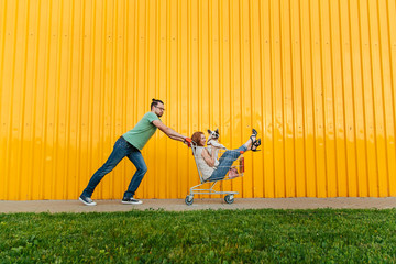 Portrait of happy young couple having fun. French dog sitting in shopping cart . Yellow supermarket...