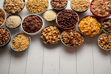 Assortment of different kinds cereals placed in ceramic bowls with cornflakes, granola, cereals and oatmeal. The concept of breakfast food. Flat lay, with copy space, top view on white wooden table.