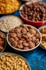 Assortment of various kinds cereals placed in ceramic bowls with cornflakes, granola, cereals and oatmeal. Flat lay, top view on blue rusty table with copy space in the middle. Selective focus.