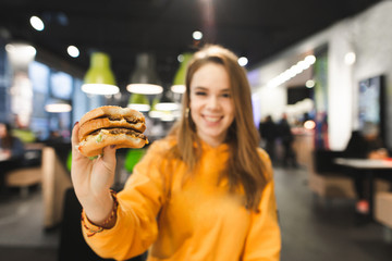 Happy girl in bright clothes keeps an appetizing big burger on the background of a fast-food restaurant and smiles. Girl student with burger in hand. Focus on the burger.