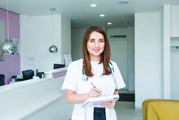 Portrait of young female doctor with note book looking at camera and smiling cheerfully while posing in receptional hall of modern clinic. Healthcare and medicine. Copy space.